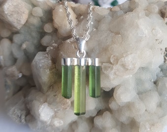 Puede incluir: Un collar con colgante de plata que presenta tres cristales de turmalina verde translúcidos dispuestos verticalmente en un marco de plata. El colgante cuelga de una cadena de plata tipo cuerda, mostrando la forma natural y alargada de las gemas.