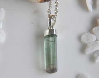 Puede incluir: Un collar con colgante que presenta un cristal de turmalina verde translúcido, largo y cilíndrico, suspendido de una anilla de metal plateado y una cadena plateada trenzada. El cristal muestra inclusiones naturales y un degradado de color.
