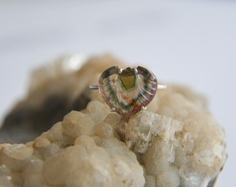 May include: A ring featuring a heart-shaped, carved, multi-coloured gemstone with bands of green, pink, and clear tones. The stone is set in a simple silver-toned metal band with four small prongs holding the gemstone securely in place.