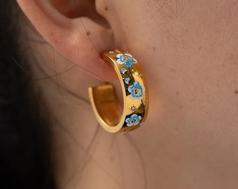 May include: A gold hoop earring featuring a polished metallic finish and a decorative pattern of small blue flowers with white centers and tiny star-like accents scattered across the outer surface of the circular band.