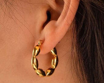 May include: A gold hoop earring featuring a series of connected, smooth, rounded, pebble-like segments. The earring has a polished, reflective metallic finish and a circular shape, secured through the earlobe with a small hinged clasp mechanism.
