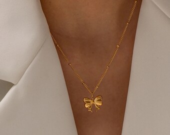 Puede incluir: Un collar de tono dorado que presenta una cadena delicada con pequeñas cuentas espaciadas uniformemente y un colgante central en forma de lazo. El colgante tiene una superficie pulida y reflectante con detalles de ranuras.