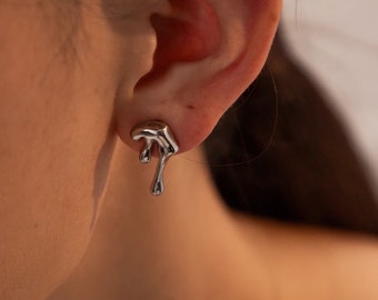 Puede incluir: Un pendiente de botón plateado con un diseño de metal fundido fluido que parece gotear desde el lóbulo de la oreja. La superficie es lisa y reflectante, captando la luz con curvas orgánicas irregulares y una extensión en forma de gota en la base.