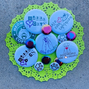 Op de afbeelding: Een verzameling ronde, lichtblauwe en metallic knopen met verschillende ontwerpen en teksten, waaronder "Hella Cats", "Cats Over Capitalism" en "Dog People Are My People". De knopen zijn gerangschikt op een neon groene, kanten onderzetter.