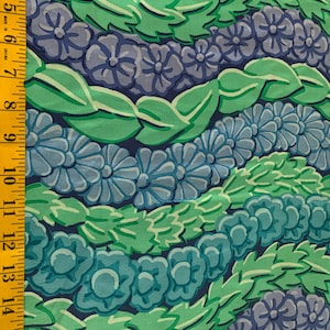 Op de afbeelding: Close-up van een stof met een herhalend bloemenpatroon. Het ontwerp toont rijen gestileerde bloemen en bladeren in tinten blauw, groen en grijs tegen een donkerblauwe achtergrond. Een gele meetlint staat aan de linkerkant.