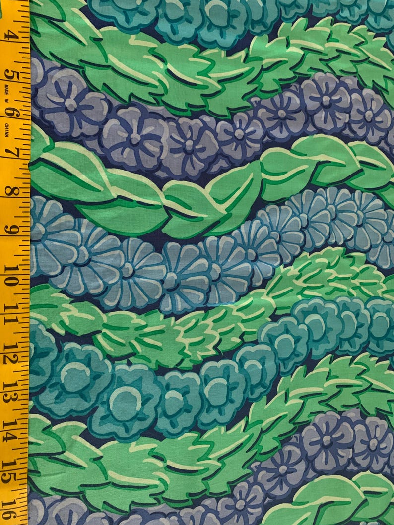 Op de afbeelding: Close-up van een stof met een herhalend bloemenpatroon. Het ontwerp toont rijen gestileerde bloemen en bladeren in tinten blauw, groen en grijs tegen een donkerblauwe achtergrond. Een gele meetlint staat aan de linkerkant.