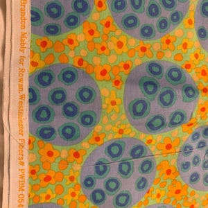 May include: A fabric featuring a light green background with a pattern of large, light purple circles containing smaller dark blue circles. Interspersed among the circles are small yellow and orange flowers. The white selvage edge reads Brandon Mably for Rowan. Westminster Fibers # PWBM-054 Pods.