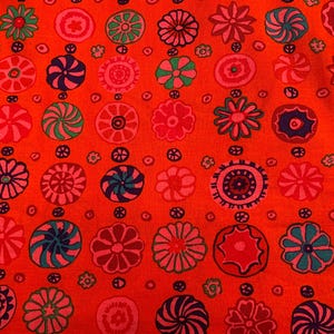 May include: Vibrant orange fabric featuring a repeating pattern of floral and circular designs in pink, blue, green, and red. The pattern is set against a solid orange background, creating a bold visual.