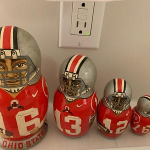 May include: Set of five nesting dolls painted to resemble football players in red and gray uniforms. Each doll features a player's face, helmet, and jersey with a different number. The largest doll has "OHIO STATE" on the base.