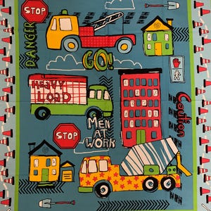 May include: A blue and green fabric with a construction theme. It features a variety of construction vehicles, including a tow truck, a dumper truck, a cement mixer, and a flatbed truck. The fabric also includes a stop sign, a danger sign, a go sign, and a men at work sign. The fabric is bordered by a row of orange traffic cones.