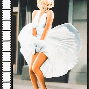 May include: A black and white photograph of a woman wearing a white dress with a pleated skirt. The woman is smiling and holding the skirt up with her hands. The photo is set against a blurred background.