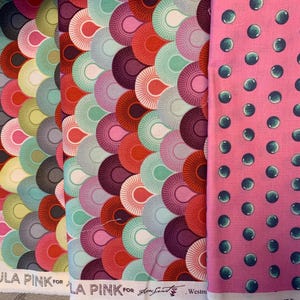 May include: Three fabric bolts with colourful patterns. Two bolts feature a repeating fan-shaped design in shades of red, pink, teal, and purple. The third bolt has a pink background with a pattern of teal bubbles. The text "TULA PINK" is visible on the bottom of the fabric.