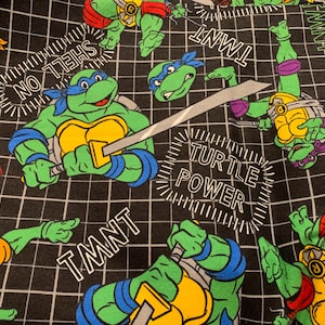 May include: A black fabric featuring a white grid pattern and a repeating print of Teenage Mutant Ninja Turtles characters in green, yellow, and blue. The design includes the text TMNT, SHELL ON, and TURTLE POWER in white and red lettering.
