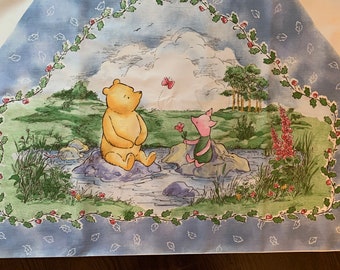 May include: A watercolor illustration of Winnie the Pooh and Piglet sitting on rocks by a stream. The background is a blue sky with white clouds and a green field. There are red berries and green leaves around the edge of the image.