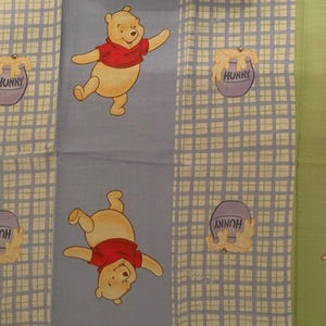Puede incluir: Tela con diseños de Winnie the Pooh y Piglet. Winnie the Pooh lleva una camisa roja y Piglet un atuendo rosa. El fondo tiene un patrón de cuadrícula con botes de miel que dicen "Hunny".