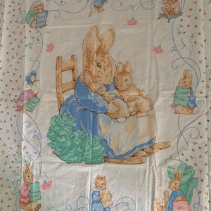 May include: A white fabric featuring illustrations from Beatrix Potter's stories. The central image shows a rabbit mother with two bunnies. Other characters include a duck, a frog, and rabbits holding signs. Pink flowers and blue vines decorate the border.