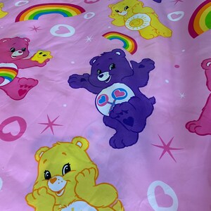 以下が含まれることがあります： ピンクの生地に、ケアベアが繰り返し描かれたパターンがあります。クマはすべて異なる色で、異なる表情をしています。生地には虹とハートも描かれています。
