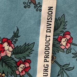 May include: Close-up of a white fabric with black text that reads "Williamsburg Product Division" and "Windham Fabrics" with a blue floral pattern on the fabric.