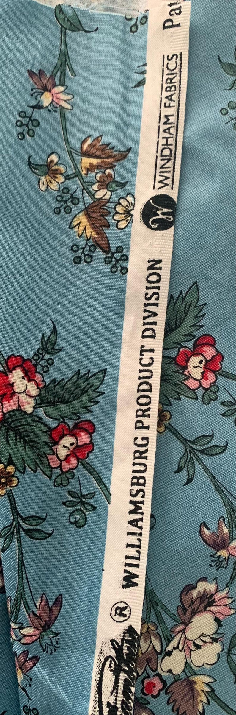 May include: Close-up of a white fabric with black text that reads "Williamsburg Product Division" and "Windham Fabrics" with a blue floral pattern on the fabric.