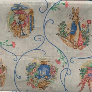 May include: White fabric with a repeating pattern of illustrations featuring characters from a children's story. The illustrations include a rabbit in a blue jacket, a rabbit sweeping, and other animal characters. The design also includes blue swirls and pink flowers.