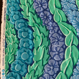 Op de afbeelding: Close-up van stof met een bloemenpatroon in blauw- en groentinten. Het ontwerp toont rijen gestileerde bloemen en bladeren tegen een donkerblauwe achtergrond. De stof is gelabeld met "Kaffe Fassett Collective" en "Serpentine".