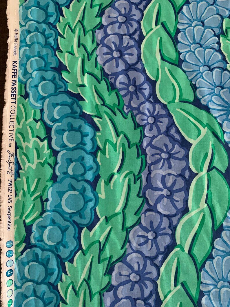 Op de afbeelding: Close-up van stof met een bloemenpatroon in blauw- en groentinten. Het ontwerp toont rijen gestileerde bloemen en bladeren tegen een donkerblauwe achtergrond. De stof is gelabeld met "Kaffe Fassett Collective" en "Serpentine".