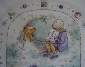 May include: A Winnie the Pooh themed plate with a light blue background. The plate features a drawing of Winnie the Pooh and a child reading a book that says "JAM" on the cover. The plate is decorated with a border of flowers, bees, and letters.