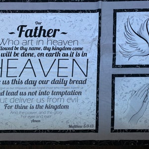 May include: A white fabric wall hanging with black trim features a framed image of a dove with an olive branch in its beak, a pair of hands in prayer, and a framed text of the Lord's Prayer. The text includes the words "Our Father" and "Matthew 6:9-13".
