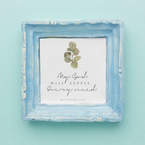 May include: A light blue frame with a white background and a watercolor illustration of a eucalyptus branch. The text "My God will supply every need" is written in black cursive font. The text "Philippians 4:19" is written in black font below the cursive text.