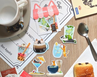 May include: A collection of colorful stickers featuring cats and drinks. The stickers are on a white surface with a brown wooden background. The stickers feature a cafe called "Calirrion Cafe" with a pink awning. The stickers also feature a cat drinking a glass of iced tea, a cat drinking a glass of coffee, a cat drinking a glass of lemonade, a cat drinking a glass of iced tea with boba, and a cat drinking a glass of matcha.