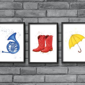 May include: Three framed watercolor prints. The first features a blue French horn, the second a pair of red cowboy boots, and the third a yellow umbrella. Each print has a white background with watercolor splatter.