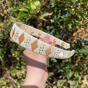 May include: A white headband with a gold and white geometric pattern. The pattern features small gold dots and a diamond shape.