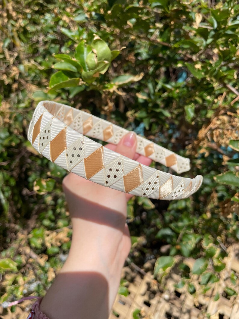 May include: A white headband with a gold and white geometric pattern. The pattern features small gold dots and a diamond shape.