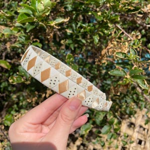 May include: A white headband with a gold and white geometric pattern. The headband is made of a soft, flexible material and is perfect for adding a touch of style to any outfit.