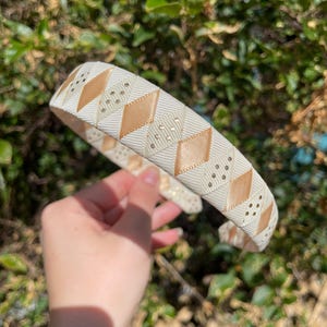 May include: A white and gold headband with a geometric pattern. The headband is made of a soft, flexible material and is perfect for adding a touch of elegance to any outfit.