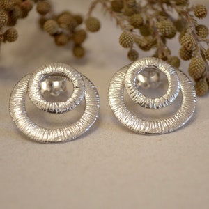 Puede incluir: Un par de pendientes de plata con un diseño de doble círculo. Los círculos son texturizados y tienen un aspecto ligeramente martillado.