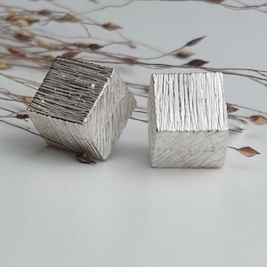 Peut inclure: Deux boucles d'oreilles carrées en argent texturé. La surface des boucles d'oreilles est rugueuse et lignée. Le fond est blanc avec des tiges de plantes séchées.