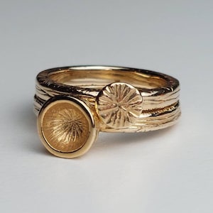 Op de afbeelding: Gouden ring met een getextureerde band en twee decoratieve elementen. Een element is een ronde schijf met een getextureerd oppervlak, de andere is een stervormige schijf met een getextureerd oppervlak.