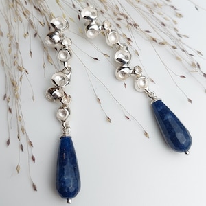 Op de afbeelding: Een paar zilveren oorbellen met een traanvormige blauwe edelsteen hanger. De oorbellen hebben een cluster van kleine, ronde zilveren kralen.