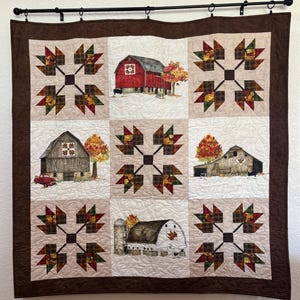 May include: A quilted wall hanging featuring a patchwork design with a brown border. The quilt showcases nine squares with alternating patterns of autumn leaves and barn scenes. The barns are red and brown, with fall foliage.