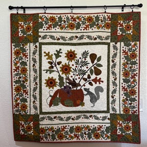 May include: A quilted wall hanging featuring a central design of sunflowers, leaves, and a squirrel in a pumpkin-shaped vase. The quilt has a white background with borders of sunflowers, acorns, and leaves in shades of orange, green, and brown.
