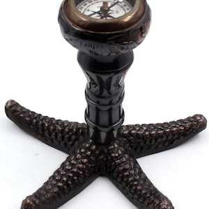 Può includere: Una bussola decorativa con finitura nera e ottone, montata su una base a forma di stella marina. Il quadrante della bussola ha uno sfondo nero con segni bianchi e un ago rosso.