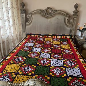 May include: A colorful quilt with a geometric pattern in red, yellow, green, and white. The quilt is on a bed with a decorative headboard. The quilt's design features repeating squares and diamond shapes.
