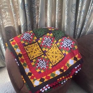 May include: A colorful, patterned quilt with a red border, yellow and green squares, and white geometric designs. The quilt is draped over a brown ottoman. The quilt's edges are trimmed with a decorative, scalloped border.