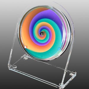 Könnte beinhalten: Ein kreisförmiges kinetisches Sandkunstobjekt mit einem leuchtenden, mehrfarbigen Spiralmuster in Violett, Türkis und Orange. Die runde Glasscheibe ist in einen klaren Acrylrahmen eingefasst, der es dem Sand im Inneren ermöglicht, sich zu bewegen.