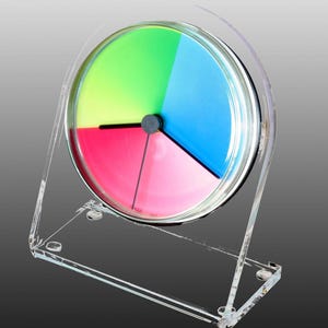 Puede incluir: Un reloj de escritorio circular con un soporte de acrílico transparente y una esfera dividida en tres segmentos de color distintos: verde brillante, azul vibrante y rosa intenso. El reloj tiene tres manecillas negras finas en el centro.