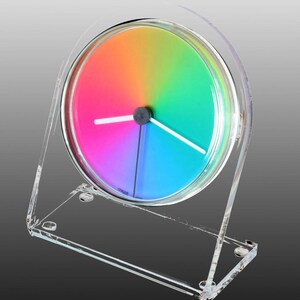 May include: A circular desk clock featuring a vibrant rainbow gradient face that transitions through colors. The clock is encased in a clear acrylic frame with a rectangular base and includes two white hands and one thin black hand at the center.