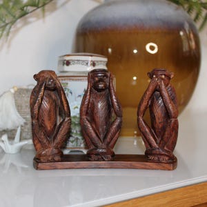 Könnte beinhalten: Drei geschnitzte Holzfiguren von Affen, die jeweils eine andere Pose einnehmen, die "Nichts sehen, nichts hören, nichts sagen" darstellt. Die Affen sind von tiefbrauner Farbe und auf einem rechteckigen Sockel montiert. Im Hintergrund ist eine dekorative Vase zu sehen.