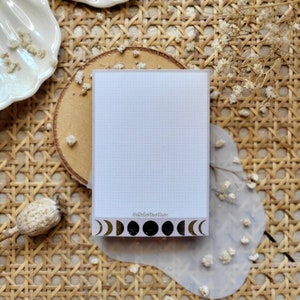 Könnte beinhalten: Ein weißer Notizblock mit einem gepunkteten Rastermuster und einem gold-schwarzen Mondphasen-Design am unteren Rand. Der Text "@atelierantan" ist auf dem Notizblock gedruckt.