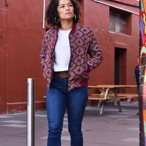 May include: A woman wearing a patterned bomber jacket with a white t-shirt and blue jeans. The jacket features a pink and red geometric design. She is also wearing a brown belt and white sneakers. The background includes a red brick wall and a graffiti-covered wall.